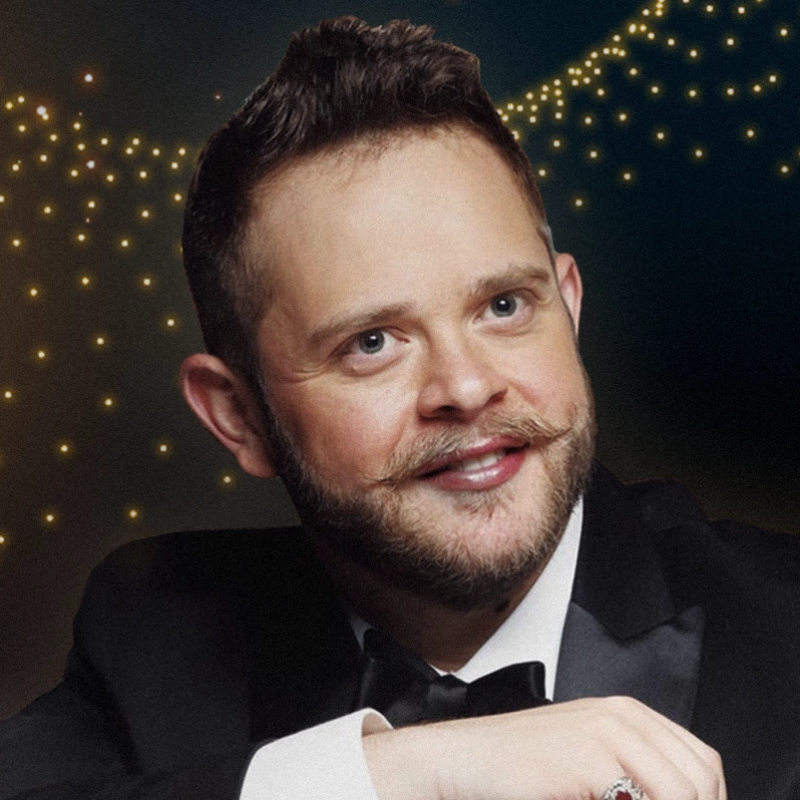 A smiling man with light skin, blue eyes, and brown hair styled up poses in a tuxedo with a bow tie. He has a styled mustache and beard. The dark background features soft, glowing string lights creating a festive, elegant atmosphere.