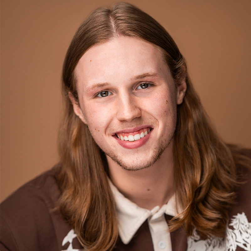 Danielle Serrano-Bremer, a young person with long, straight, light brown hair and fair skin, smiles warmly. They wear a brown shirt with a white floral pattern over a cream-colored collared shirt against a plain, warm beige background.