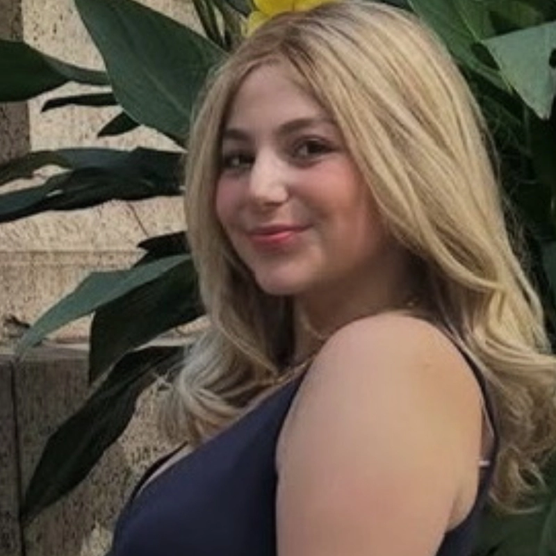 Talia Brandwein, a young woman with long, wavy blonde hair, smiles slightly while looking at the camera. She wears a sleeveless dark dress and stands in front of large green leaves and a stone wall, sunlight brightening the scene.