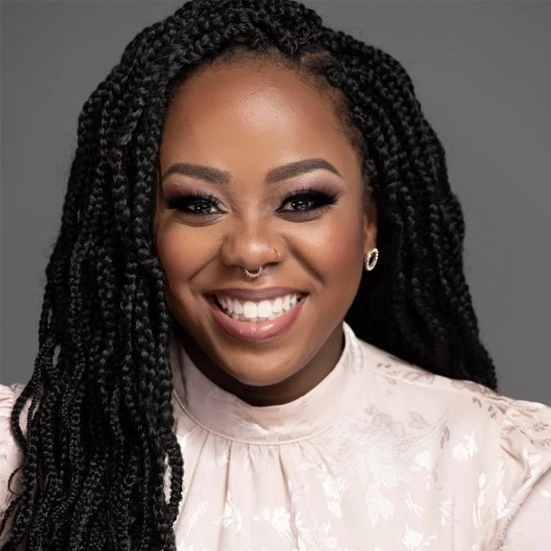 A smiling woman with long black box braids wears a pale pink, high-neck blouse with a subtle floral pattern. Breanne Sensening has glowing skin, defined makeup, hoop earrings, and a nose ring against a plain gray background.