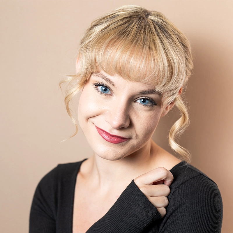 A young woman with light skin, bright blue eyes, and short blonde hair with bangs smiles softly. Caroline Bowers wears a black top and stands against a beige background, her hand gently touching her chest as a loose curl frames her face.