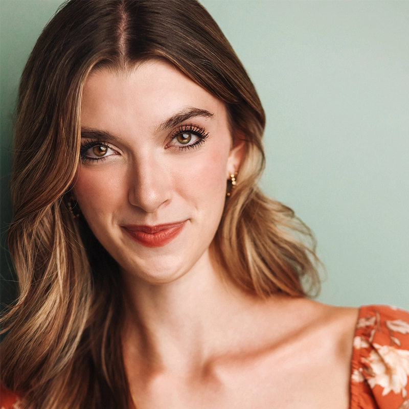 Kenzie Woolridge, a young woman with long, wavy brown hair and hazel eyes, smiles gently in front of a light green background. She wears small gold hoop earrings and a floral-patterned orange dress with a square neckline. Her makeup is natural and subtle.