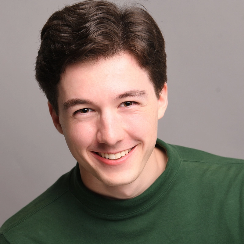 Jeremiah Burch, a young man with fair skin and short, dark brown hair, smiles warmly at the camera. He is wearing a green crew neck shirt and is posed against a plain, light gray background, his head slightly tilted to the right.