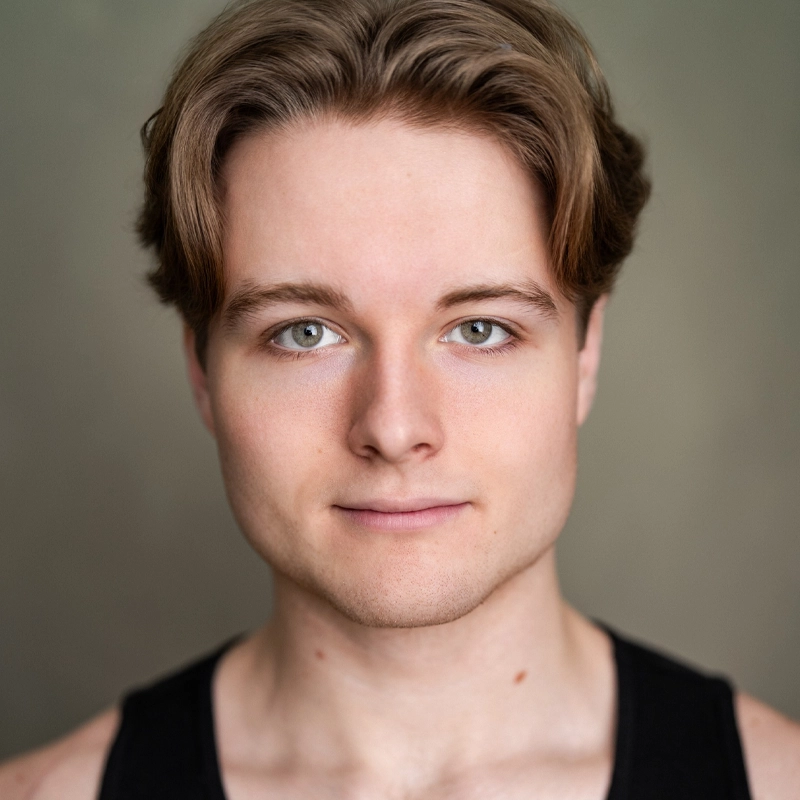 A young person with fair skin, light brown hair parted in the middle, and green eyes faces the camera directly. Mikel Campbell wears a black tank top and has a calm, neutral expression. The softly blurred background is in shades of green and brown.
