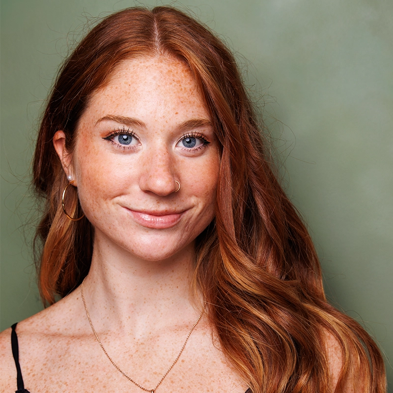 Caroline Bowers, a young woman with long, wavy red hair and fair, freckled skin, smiles gently. She has blue eyes, a nose ring, hoop earrings, and wears a thin gold necklace and a black strap top against a soft green background.