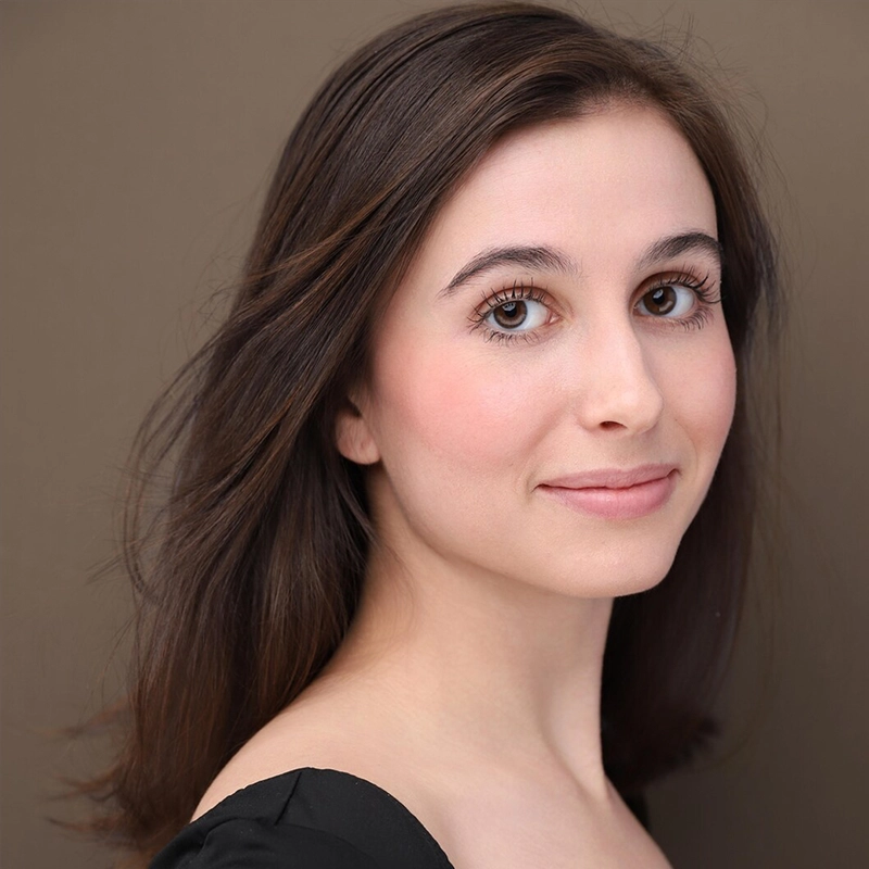 A young woman with long brown hair and fair skin smiles softly. She has brown eyes, subtle makeup, and wears a black top. The background is plain and brown, and her hair flows gently behind her as she gazes slightly past the camera.