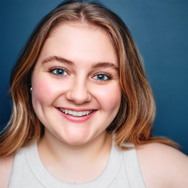 Caroline Weston, a young woman with long, light brown hair, smiles warmly at the camera. She has blue eyes, prominent dimples, and is wearing a sleeveless light beige top against a solid dark blue background.