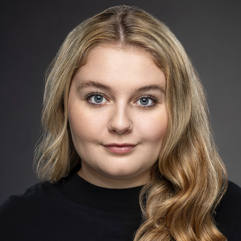 Caroline Weston, a young woman with long, wavy blonde hair and blue eyes, faces the camera against a neutral gray background. She wears a black top and has a calm, confident expression with a faint smile, her features softly highlighted by even lighting.