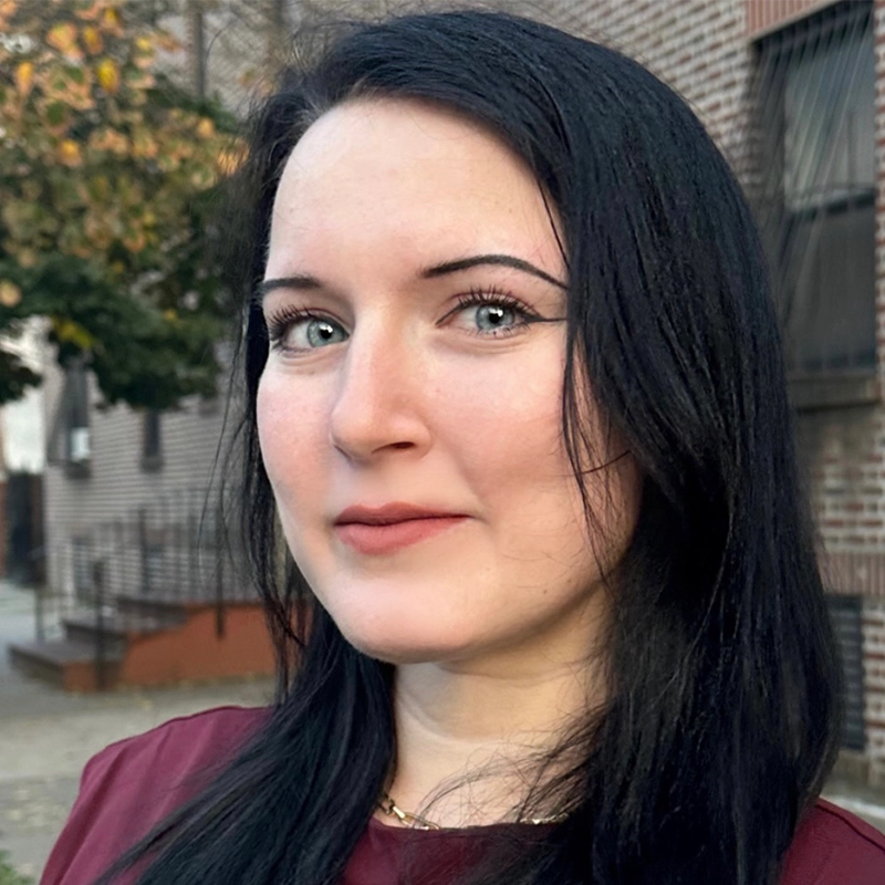 A woman with fair skin and long straight black hair stands outdoors in front of a brick building. She has light-colored eyes, arched eyebrows, and wears natural makeup with a berry-toned lipstick. She is dressed in a maroon top.