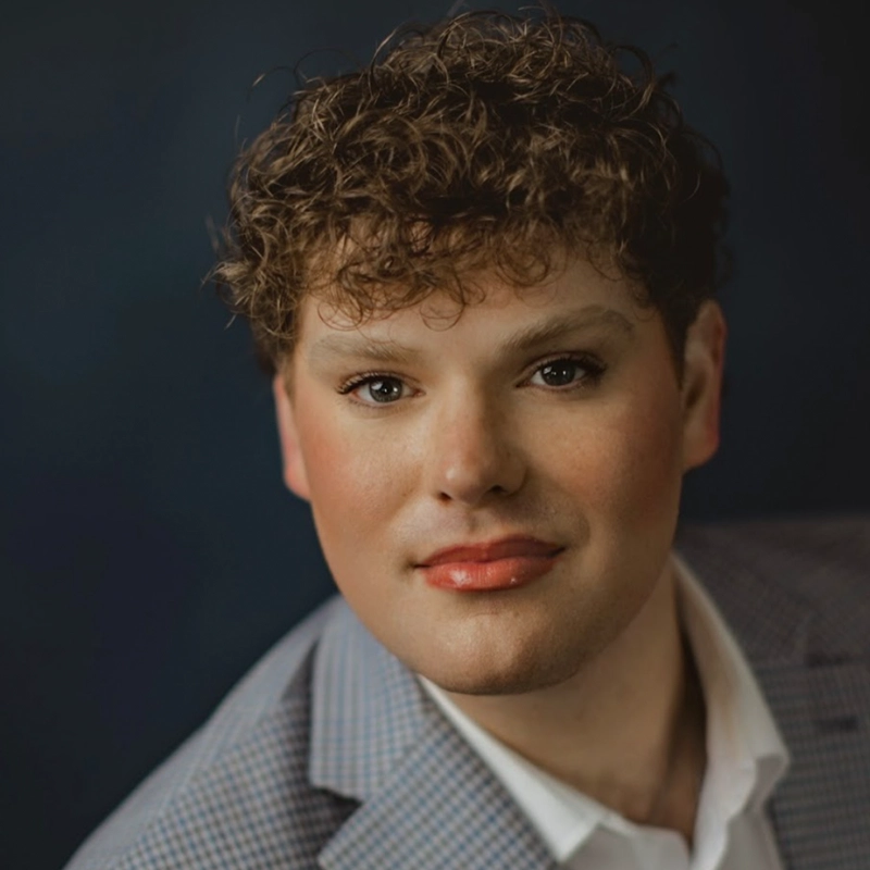 A person with curly brown hair and light skin is wearing makeup, including lipstick and eye makeup. They are dressed in a light gray plaid blazer over a white shirt and are posed against a dark blue background, looking directly at the camera with a soft expression.