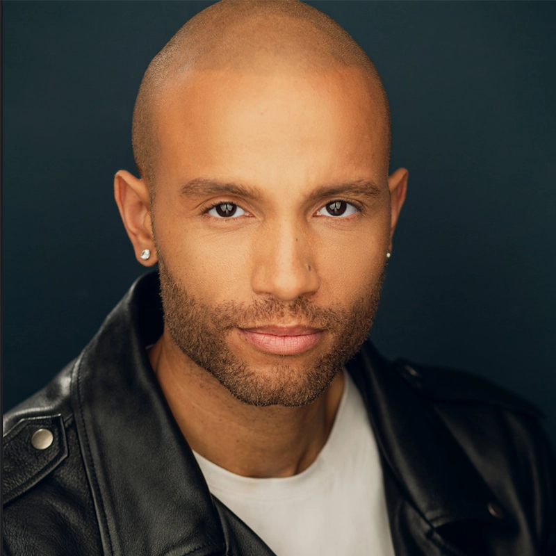 Christian Brailsford, a bald man with medium brown skin, trimmed beard, and diamond stud earrings, wears a black leather jacket over a white shirt. He faces forward with a slight smile against a dark, plain background.