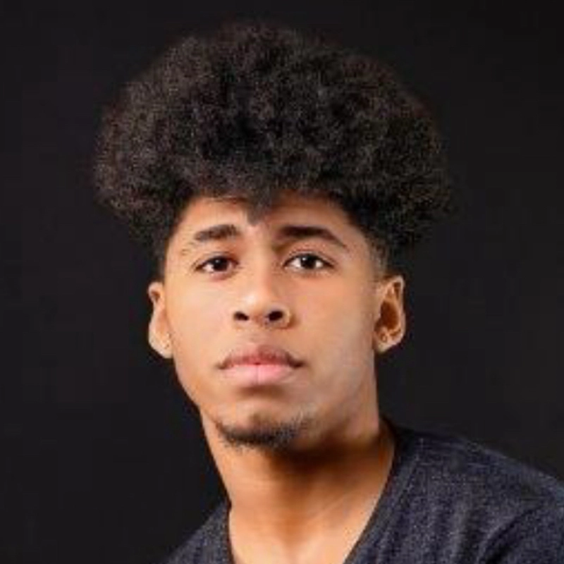A young man with medium brown skin and short facial hair looks at the camera. He has voluminous, curly dark hair, similar to Kenzie Woolridge’s signature style, and wears a simple dark gray shirt against a plain black background.