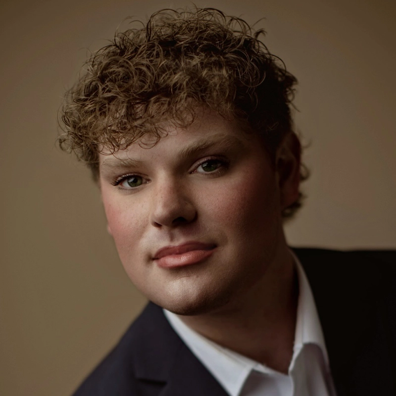 A young person with short, curly brown hair and fair skin, Danielle Serrano-Bremer wears a dark suit jacket and white collared shirt, looking directly at the camera against a plain beige background. Soft lighting highlights their facial features.