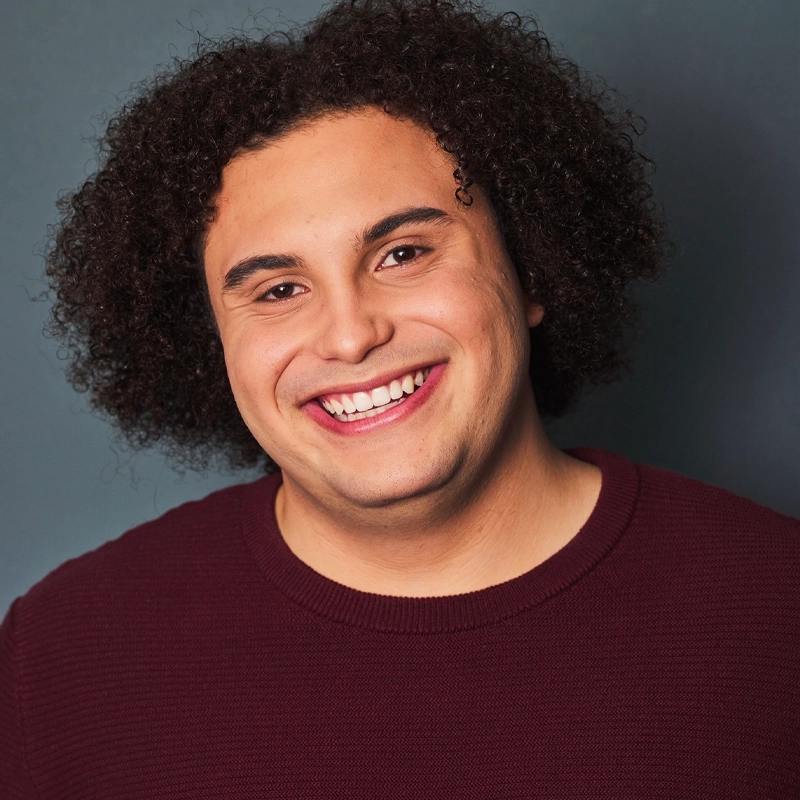 A young man with curly dark hair, Xavier Cornell, smiles broadly at the camera. He is wearing a textured, burgundy sweater and is posed against a plain, dark gray background. Soft lighting highlights his cheerful expression and smooth complexion.