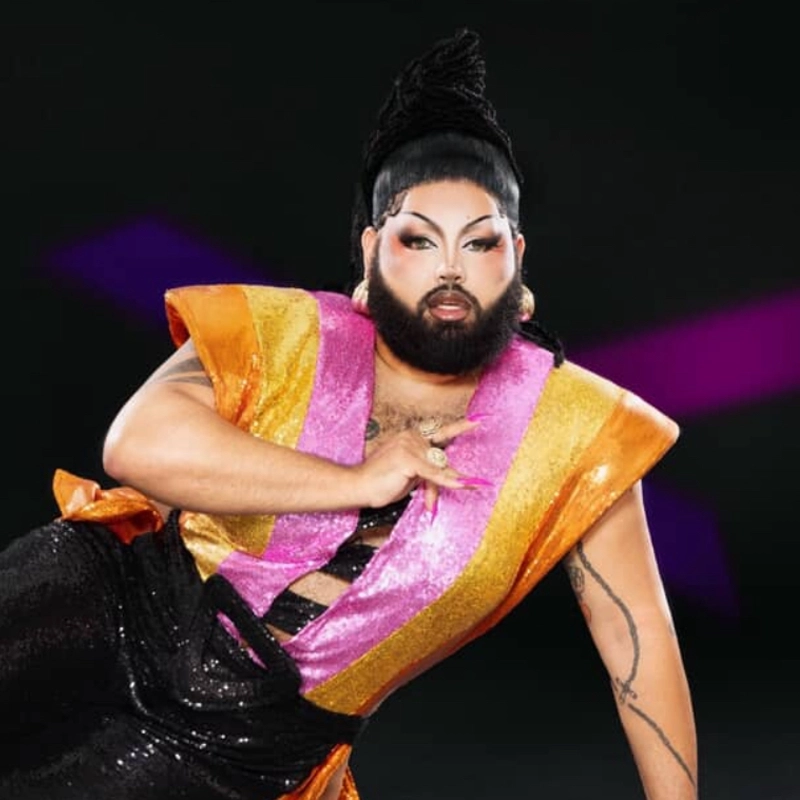 A person poses confidently against a dark background, wearing bold makeup, a dramatic black wig styled upwards, and a colorful outfit with orange, pink, and yellow shiny panels. The look is glamorous, with sequins and dramatic, arched eyebrows.
