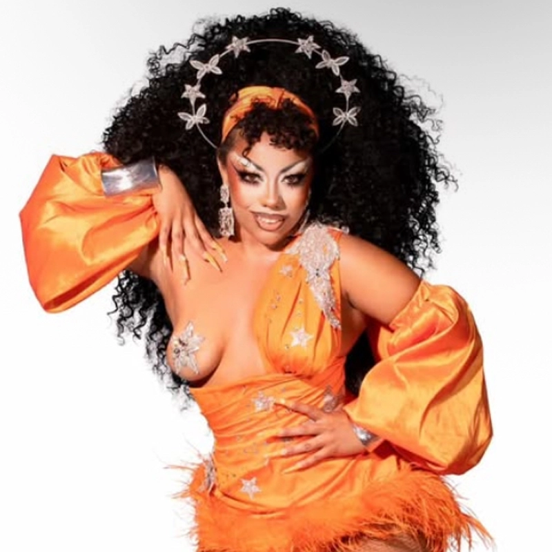 A person with voluminous curly hair wears an orange satin dress with one shoulder exposed, adorned with silver star accents and feathers. They have dramatic makeup, long nails, and a headpiece of silver stars forming a halo. The background is white.