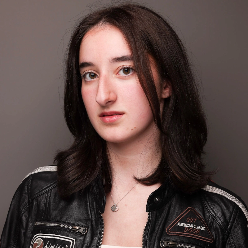 Sophia D'Erasmo, a young woman with shoulder-length dark brown hair, wears a black leather jacket adorned with patches and a simple necklace. She gazes directly at the camera, her expression neutral, posed against a plain gray background.