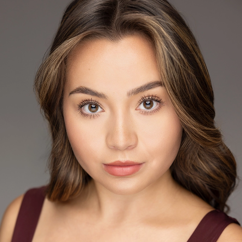 Danielle Serrano-Bremer, a young woman with wavy brown hair and light highlights, looks directly at the camera. She has brown eyes, defined eyebrows, natural makeup with nude lipstick, and wears a sleeveless maroon top against a gray background.
