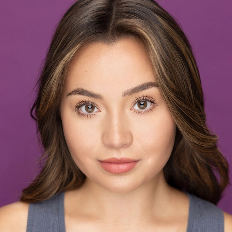 Kenzie Woolridge, a young woman with medium-length brown hair and brown eyes, looks directly at the camera in a gray top. Her expression is neutral with a slight smile. The solid purple background and even lighting highlight her facial features.