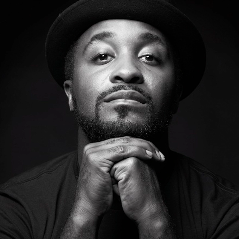 Black and white portrait of Rickey Tripp, a bearded man in a hat and dark shirt. He gazes confidently at the camera, resting his chin on clasped hands. Dramatic lighting highlights his striking facial features.