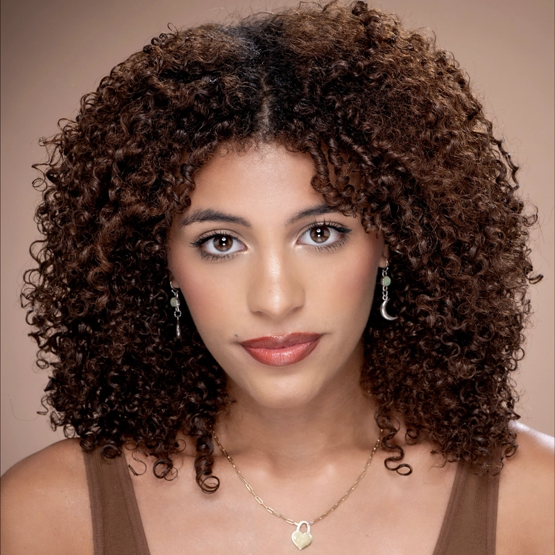 A young woman with medium-toned skin and dark, tightly curled hair poses against a brown background. Zayna Darres wears a brown top, gold earrings, a heart pendant necklace, and natural makeup with red lipstick as she looks at the camera.
