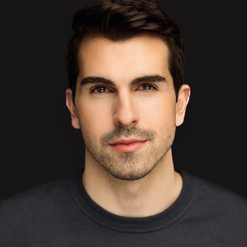David Assetta, a young man with short dark hair and neatly trimmed facial hair, looks directly at the camera. He wears a plain dark shirt and has a neutral expression. The solid black background emphasizes his face.