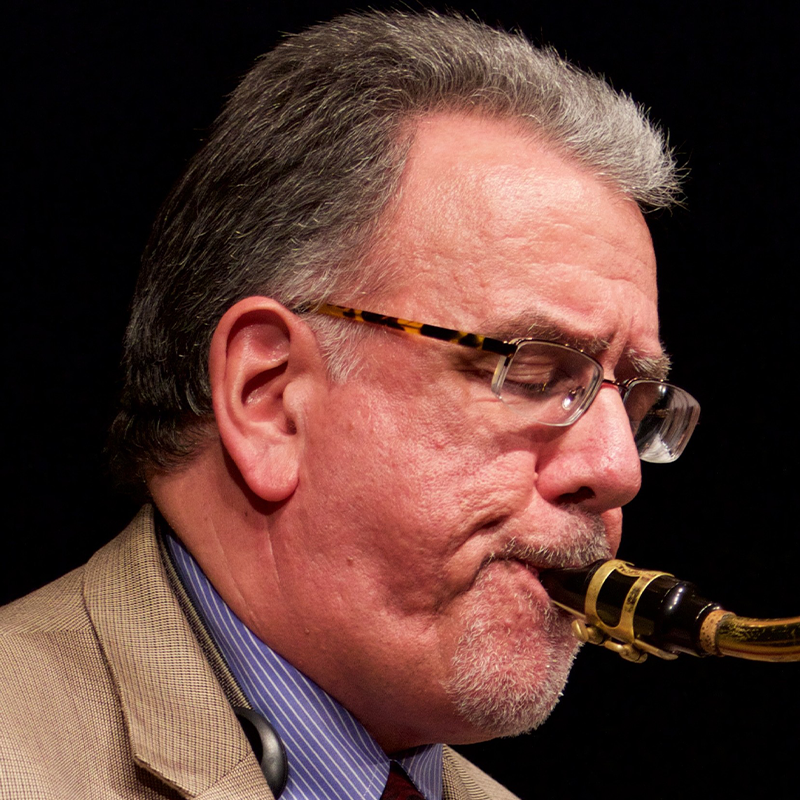 Close-up of David Demsey, an older man with glasses and gray hair in a tan suit and blue-striped shirt, playing a saxophone. His eyes are closed in concentration, set against a dark background that highlights him and the instrument.