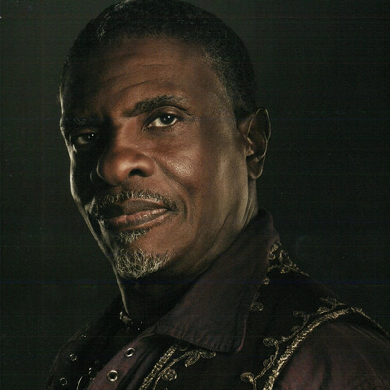 A middle-aged man with short, curly black hair and a neatly trimmed goatee gazes confidently at the camera. He wears a deep purple shirt with ornate, gold-embroidered patterns, against a dark background with dramatic lighting on his face.