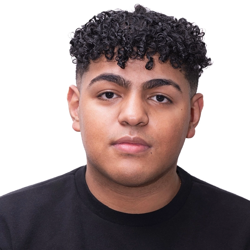 A young person with short, dark, tightly curled hair and thick eyebrows looks directly at the camera with a neutral expression. Lauren Torey is wearing a black crew-neck shirt and is posed against a plain white background.