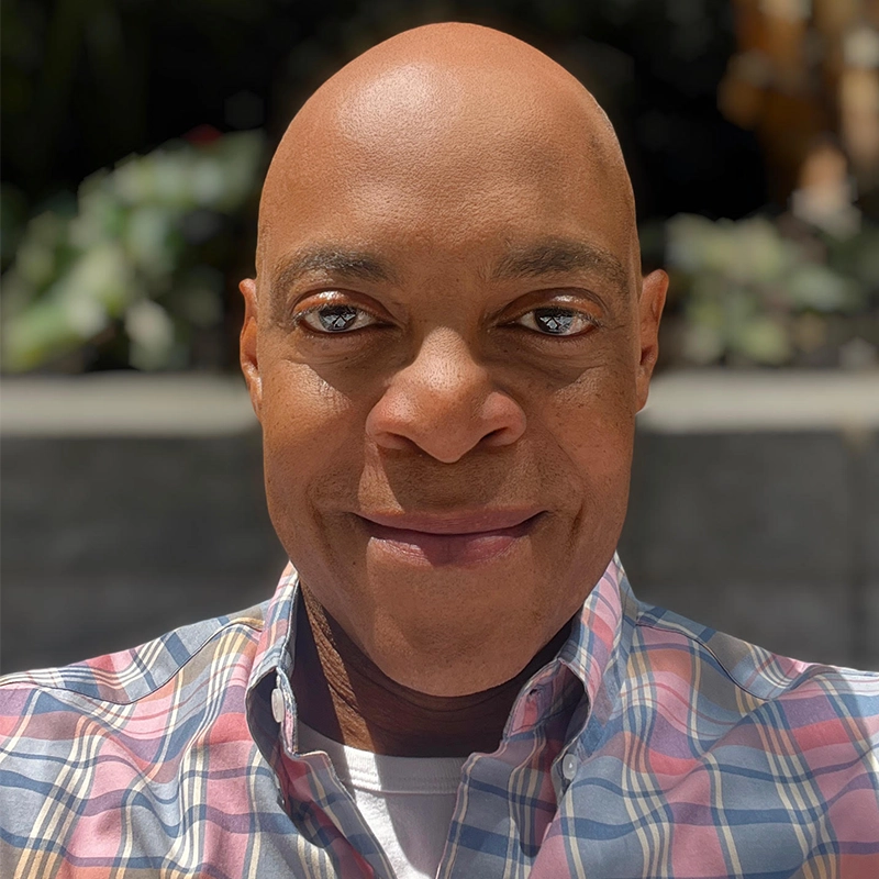 A bald man with brown skin smiles slightly while wearing a plaid button-up shirt. The background is blurred greenery and sunlight, highlighting his face. The photo is well-lit, capturing his friendly expression in a close-up, centered selfie.