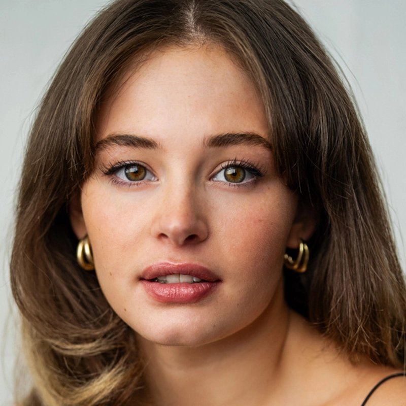Sadie Dickerson, a young woman with light skin, long brown hair, and hazel eyes, looks directly at the camera. She has natural makeup, full lips, and gold hoop earrings. The softly blurred background draws attention to her face.