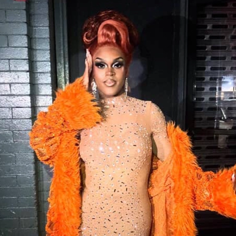 Blossom Dior stands confidently in glamorous drag, wearing a sparkling nude, body-hugging dress adorned with rhinestones and a bright orange furry coat. Dramatic makeup, jeweled earrings, and voluminous styled red hair complete the dazzling look.