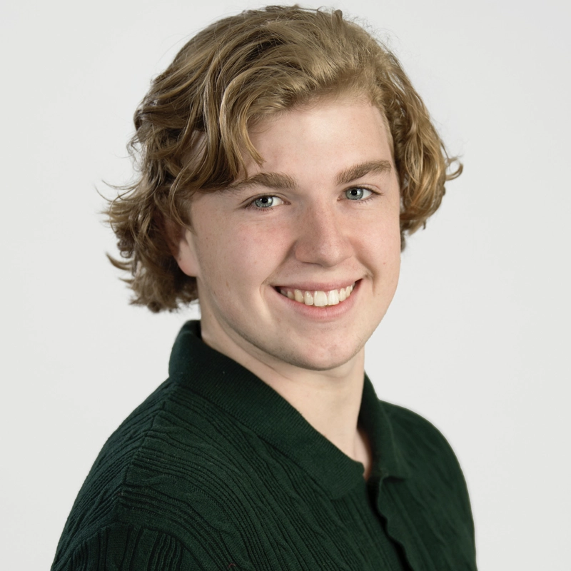 A young person with wavy blond hair and fair skin smiles at the camera. They are wearing a dark green, textured collared shirt. The background is plain white, and the person is posed at a slight angle, facing forward.