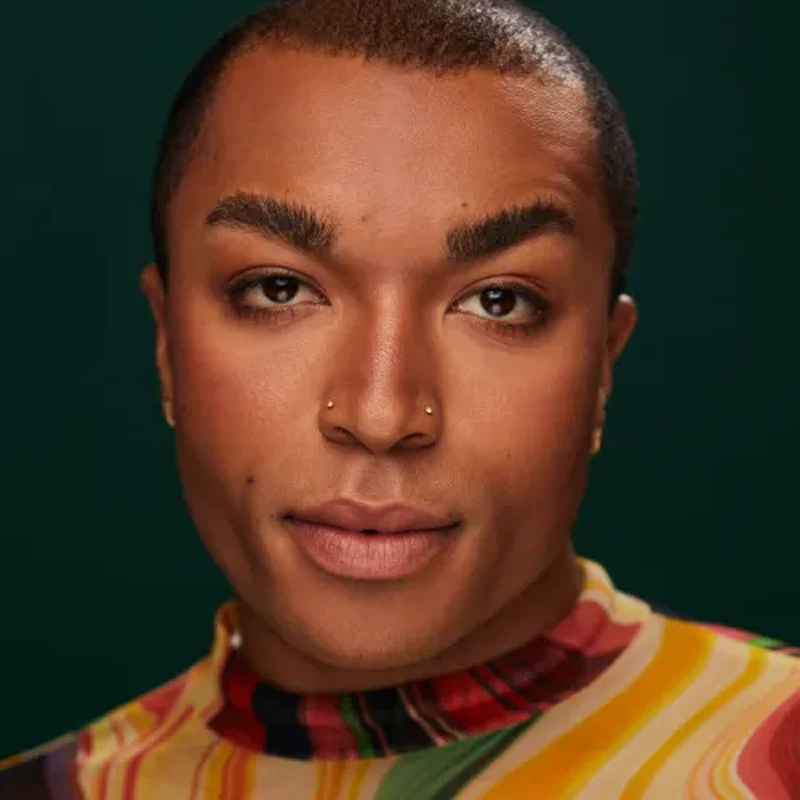 A person with short hair, smooth brown skin, subtle makeup, and double nose piercings looks confidently at the camera. They wear a high-neck, multicolored patterned top against a deep green background.