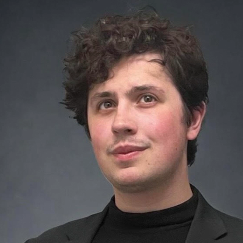 A young person with fair skin, short curly brown hair, and light facial hair is wearing a black turtleneck and dark blazer. They are looking slightly upward and to the left, set against a plain dark gray background.