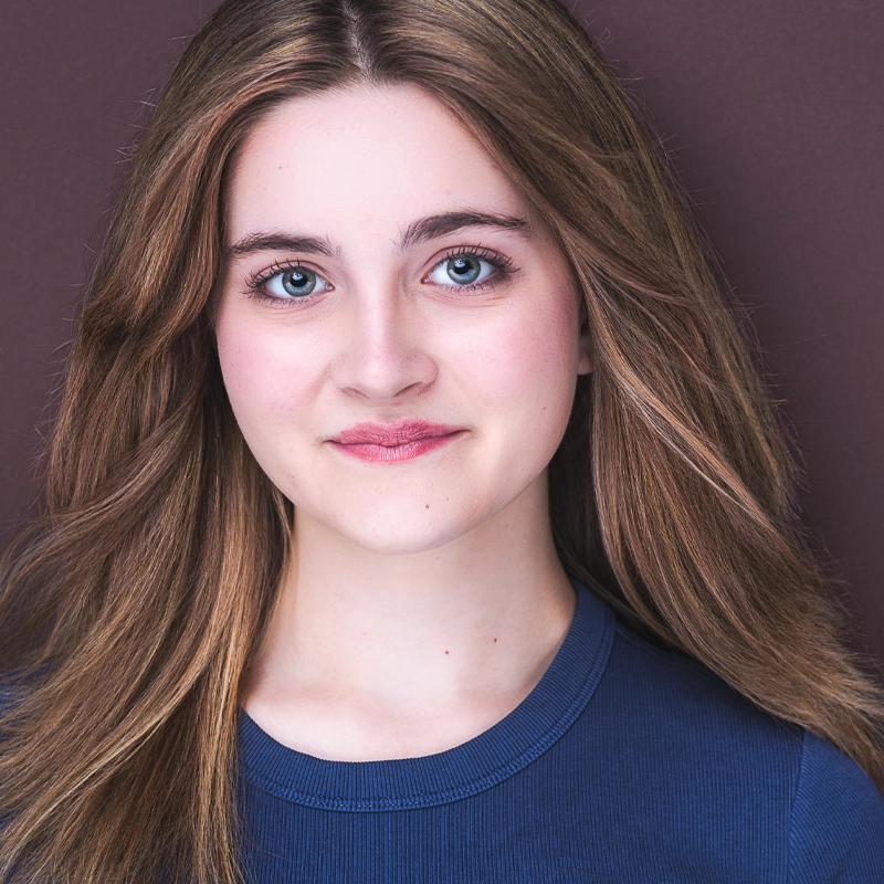 A young woman with long, light brown hair and blue eyes smiles softly at the camera. She wears a blue top and poses in front of a plain, muted brown background. Her hair is parted slightly to the side and she has a natural makeup look.