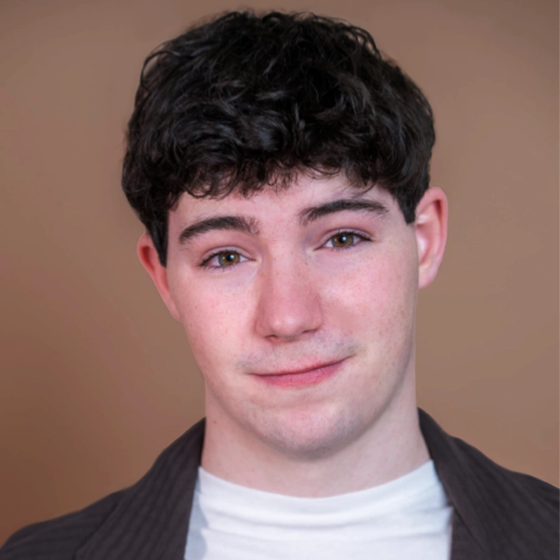 A young person with short, curly dark hair and fair skin poses in front of a plain, beige background. They wear a white t-shirt under a dark, open jacket and have a slight, friendly smile, looking directly at the camera.