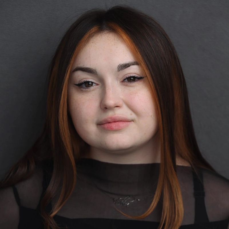 A young woman with long straight dark hair and copper highlights gazes at the camera in this striking portrait by Michael Mariniello. She has winged eyeliner, full lips, and a faint smile. She wears a sheer black top over a black shirt, and the background is plain, medium gray.