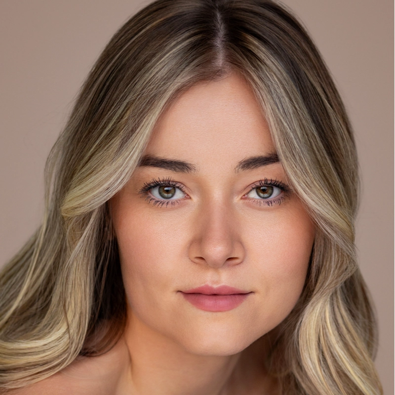 A young woman with fair skin and long, blonde, wavy hair is facing forward. She has green eyes, subtle makeup, and a neutral expression. The background is plain and beige, giving the portrait a soft, natural look in the signature style of photographer Michael Mariniello.