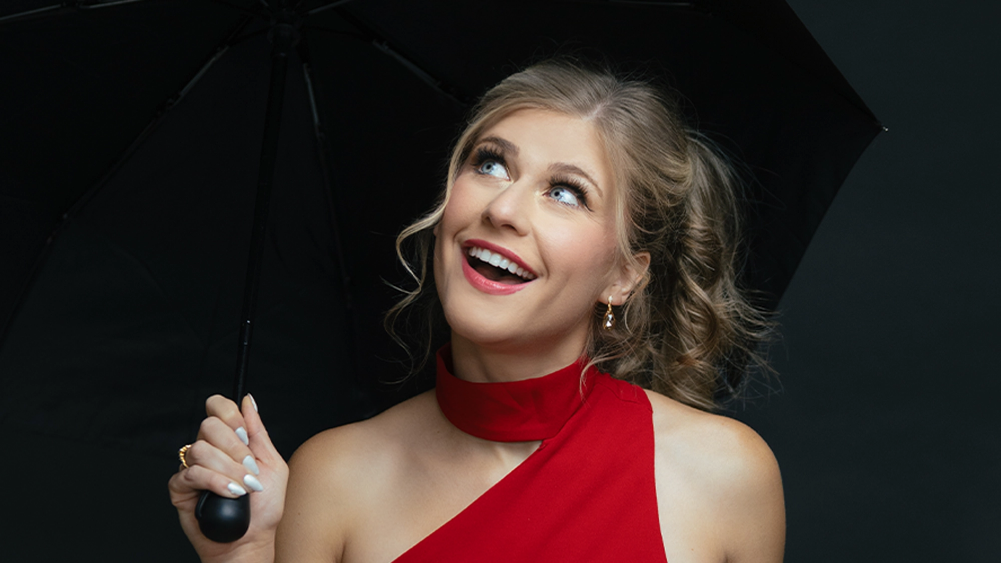 Ellen Grace Diehl site image 26 A smiling woman with wavy blonde hair holds a black umbrella, evoking Singin’ in a Rainstorm. She wears a one-shoulder red dress, gold earrings, and has manicured white nails. Her expression is happy as she looks upward against the dark background.