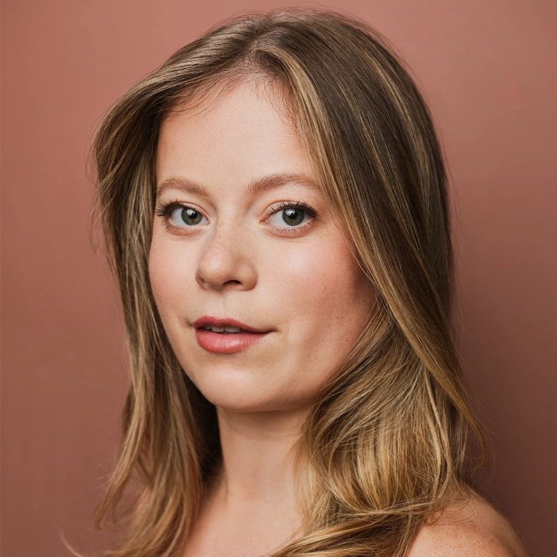 A woman with fair skin and long, straight, blonde hair poses in front of a plain, muted pink background, gazing directly at the camera with a neutral expression. She wears natural makeup and her hair is parted slightly to the side, embodying the refined yet understated elegance often captured in Michael Mariniello’s portraits.