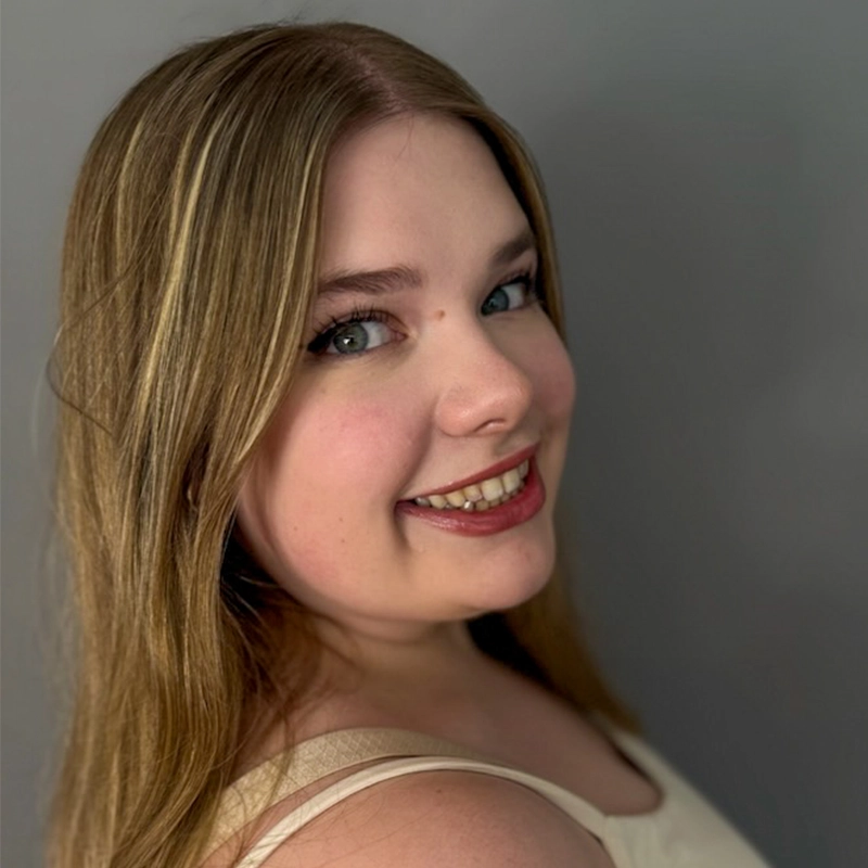 A young woman with long, light brown hair smiles at the camera. She wears a sleeveless, light-colored top and has subtle makeup with winged eyeliner and pink lipstick. The background is plain gray, and she is positioned slightly turned to her left.