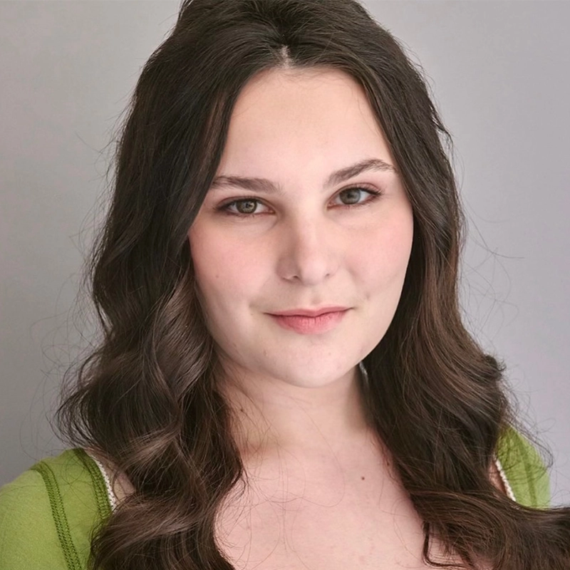 Rynn Stephenson, a young woman with long, wavy brown hair, wears a light green top and looks at the camera with a slight smile. The plain light gray background adds to the professional and clean appearance.