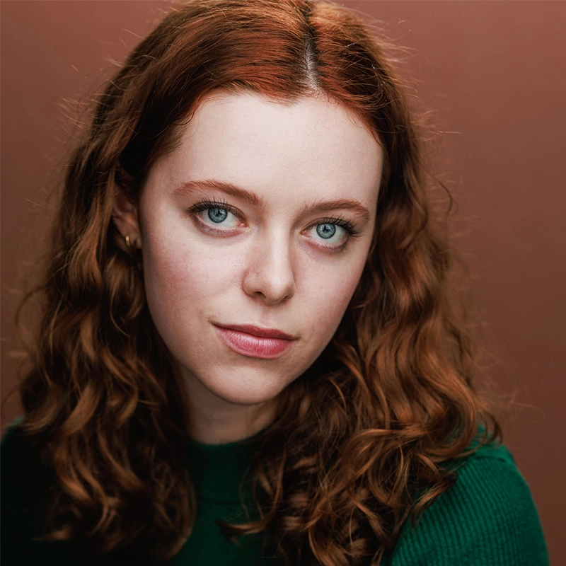 A young woman with fair skin, wavy red hair, and blue eyes looks directly at the camera with a neutral expression. She is wearing a green sweater and simple earrings. The background is a solid, warm brown color—an image reminiscent of Mickey Wirtz’s style.
