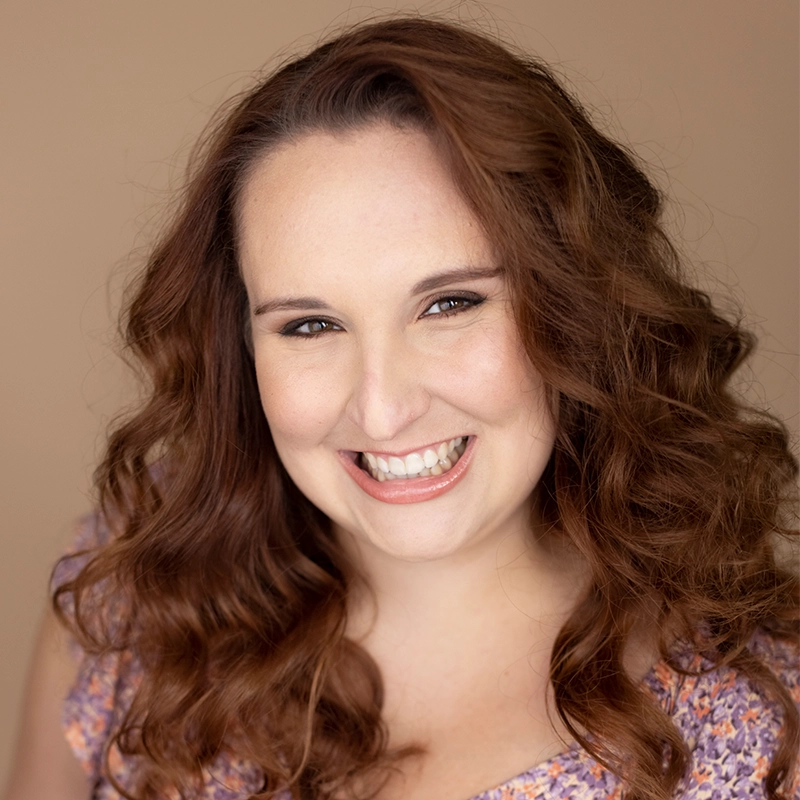 Caroline Weston, a smiling woman with long, wavy auburn hair and fair skin, wears a light purple floral top. Photographed against a simple beige background, her cheerful expression is warm and welcoming.
