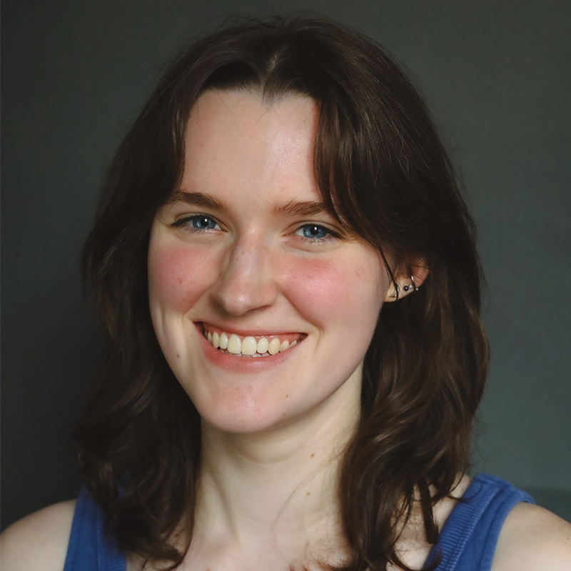 Caroline Weston, a young woman with fair skin, shoulder-length brown hair, and blue eyes, smiles warmly at the camera. She is wearing a blue sleeveless top and small earrings against a solid, dark gray background.