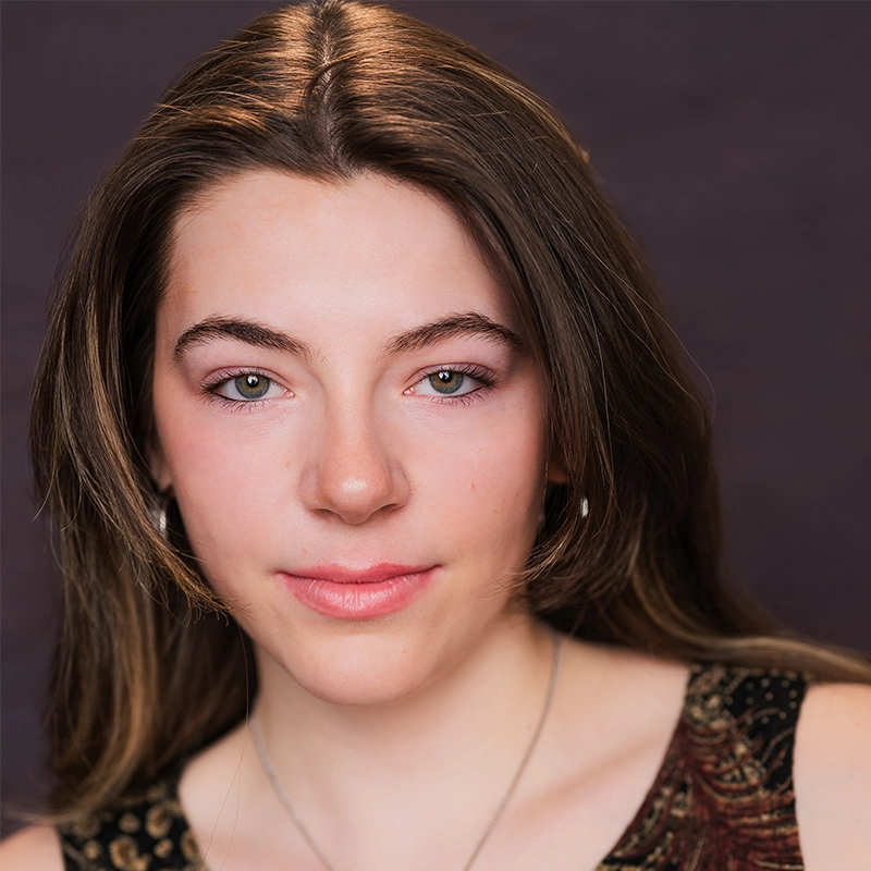 Rynn Stephenson, a young woman with fair skin and long brown hair, looks directly at the camera. She wears light pink makeup, subtle eyeliner, a sleeveless patterned top, silver hoop earrings, and a necklace against a dark background.