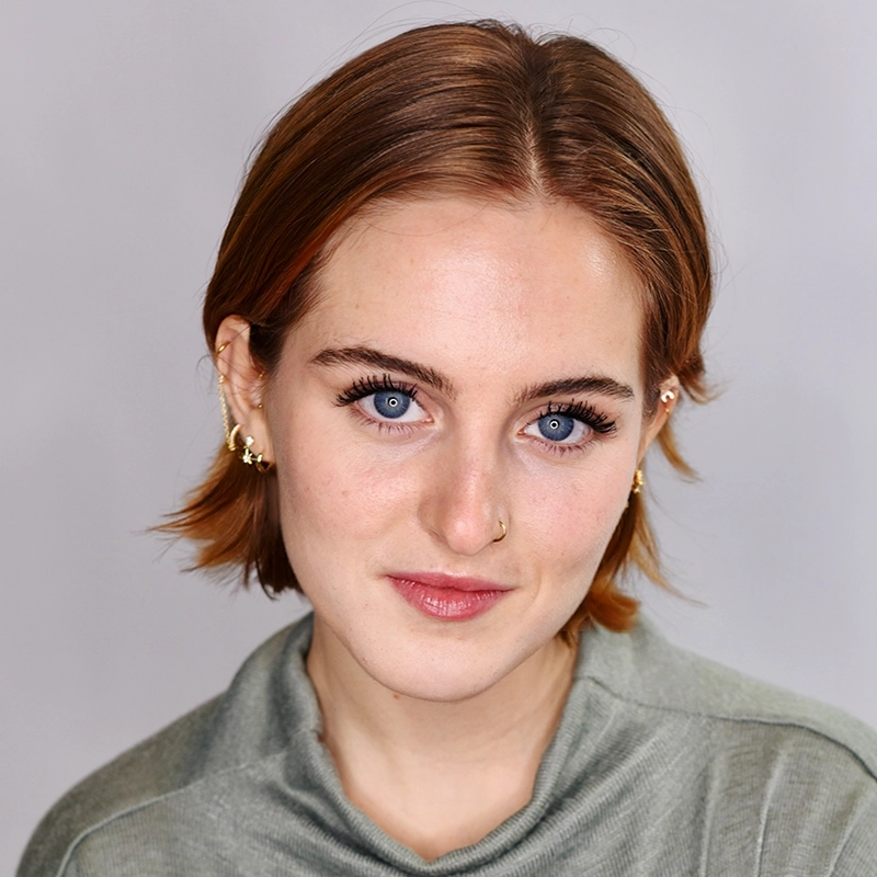 A young person with short, light brown hair and blue eyes looks at the camera, smiling slightly. Rynn Stephenson wears gold earrings, a nose ring, and a light greenish-gray top. The background is plain and light-colored.