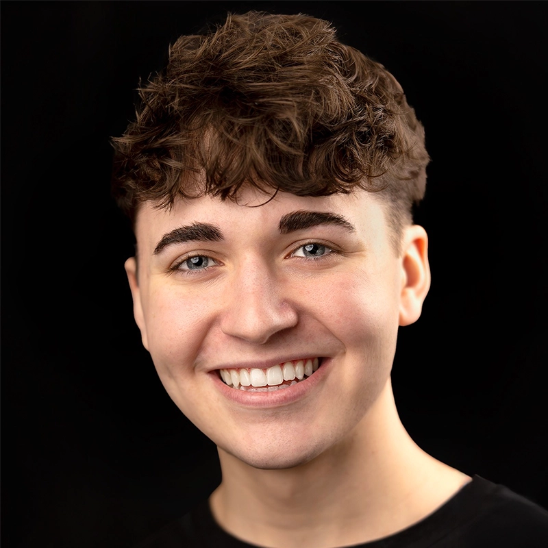 Kenzie Woolridge, a young person with short, curly brown hair and blue eyes, smiles warmly at the camera. They have fair skin, neatly groomed eyebrows, and wear a black shirt against a solid black background that highlights their face.