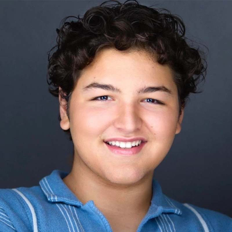 A teenage boy with curly dark hair and light skin smiles at the camera. He wears a blue collared shirt with white stripes. The background is plain and dark, drawing focus to his friendly expression and youthful features.